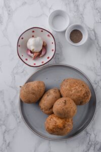 Russet potatoes on a plate, a bowl of garlic bulbs, and small bowls of salt and pepper.