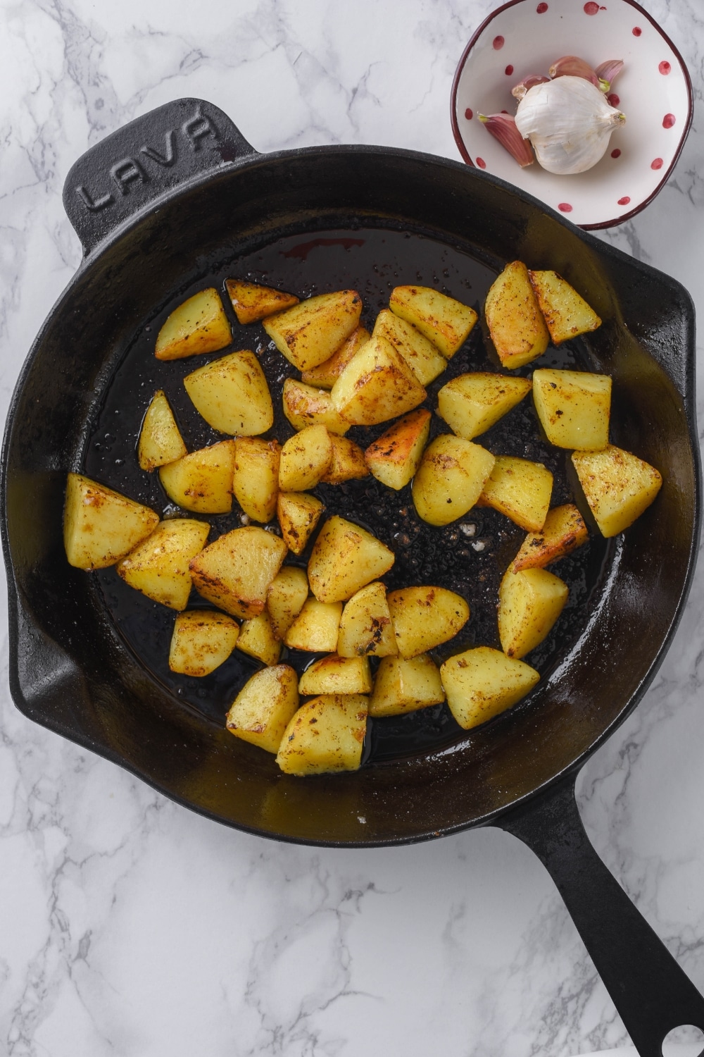 Chopped potatoes sauteeing in a cast iron skillet next to a small bowl of garlic bulbs.