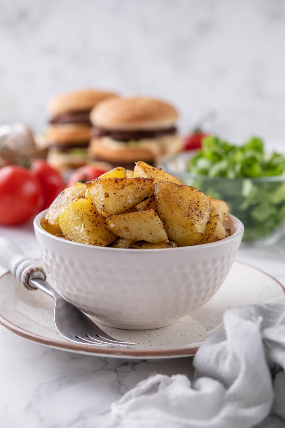 Sauteed chopped potatoes in a white bowl, set on a plate with a fork. Behind is a bowl of salad and some stacked hamburgers.