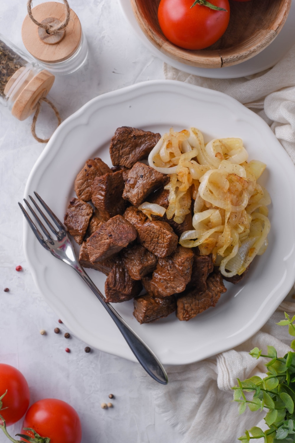 Top view of steak tips served with caramelized onions on a white plate with a fork. Surrounding it is a bowl of tomatoes and a pair of salt and pepper shakers.