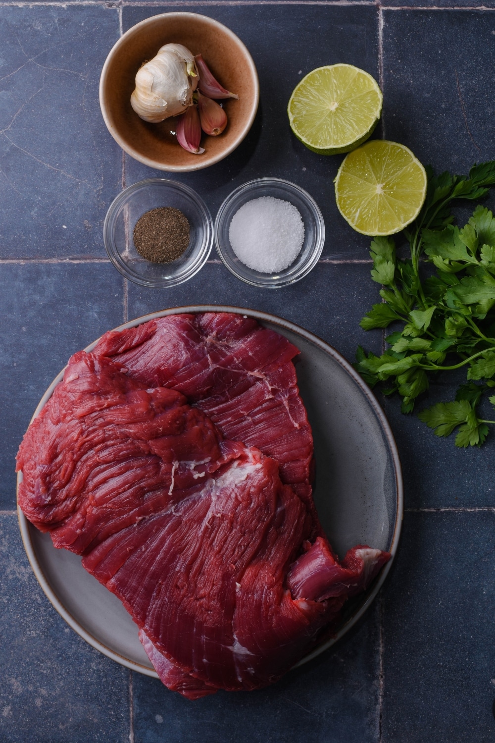 Raw skirt steak on a plate. Next to it are small bowls of salt, pepper, garlic cloves, and a halved lime.