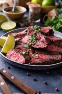 Skirt steak slices on a plate topped with chimichurri and served with lime wedges.