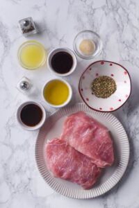 A plate of raw thick cut pork chops, small bowls of Italian herbs, olive oil, soy sauce, lemon juice, Worcestershire sauce, and garlic powder, and salt and pepper shakers.