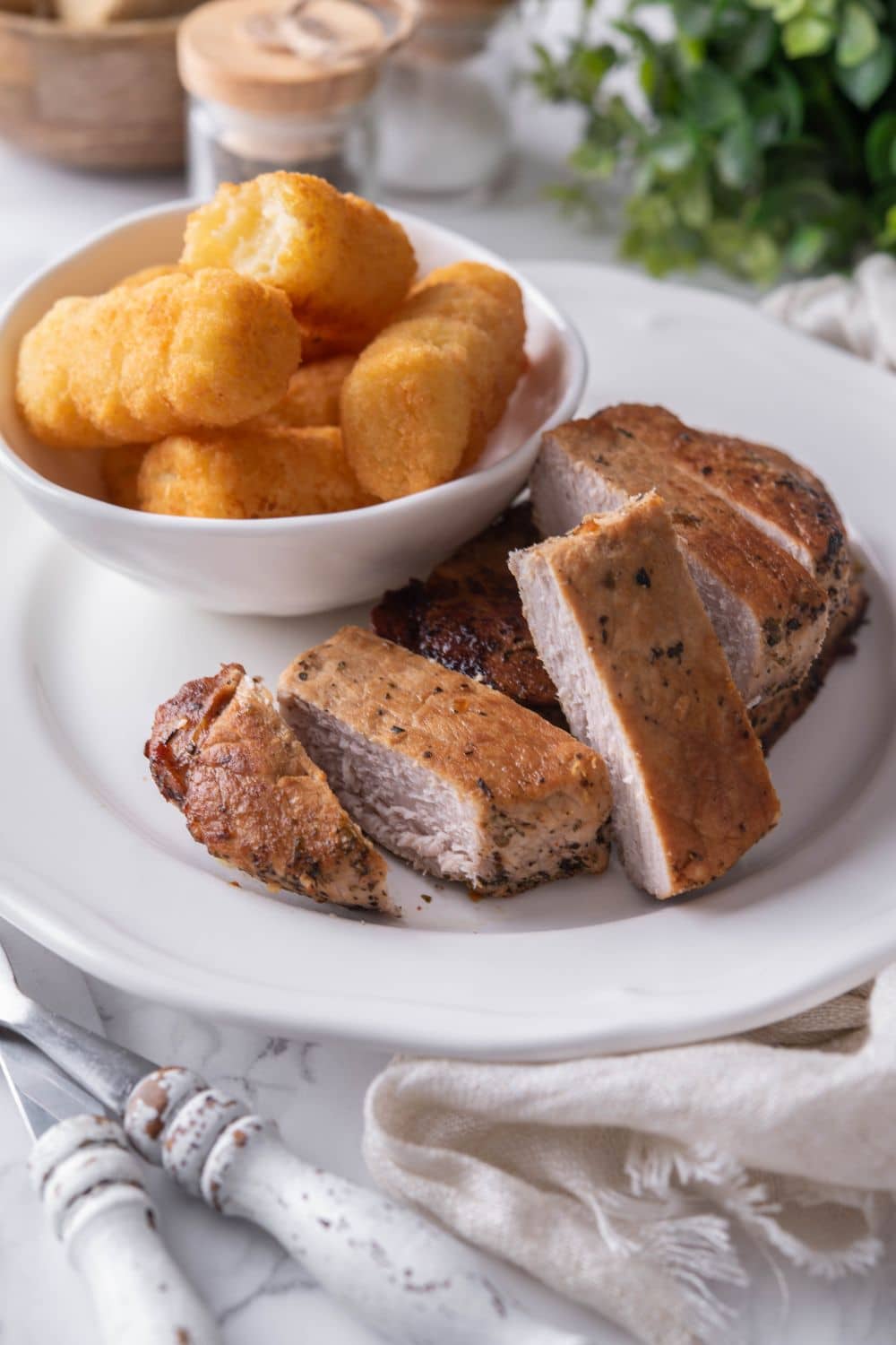 Sliced thick cut pork chops on a plate with a side of tater tots in a small bowl.