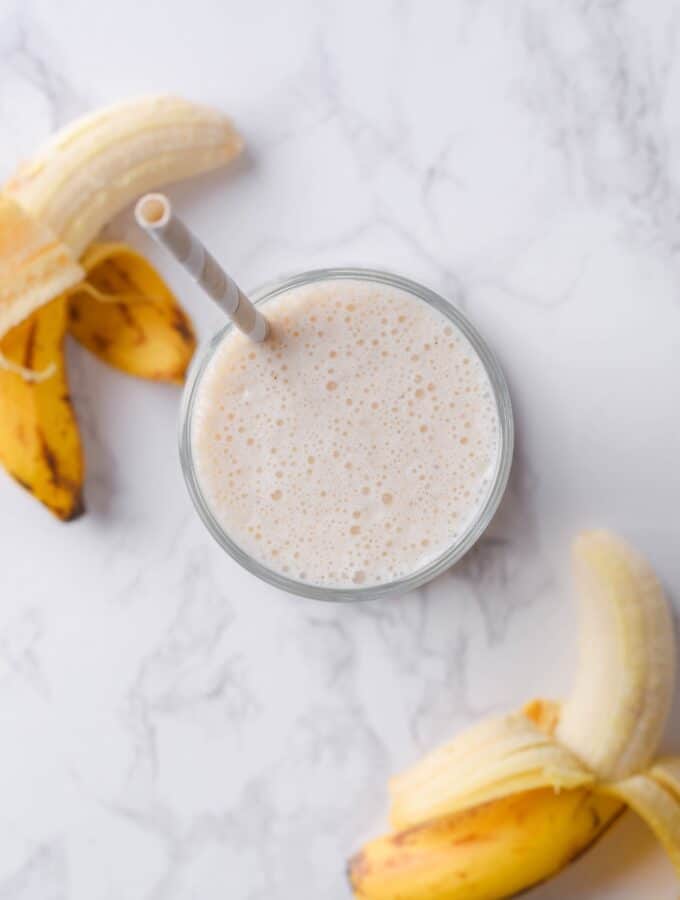 A banana protein shake in a glass on a counter with two bananas next to it.