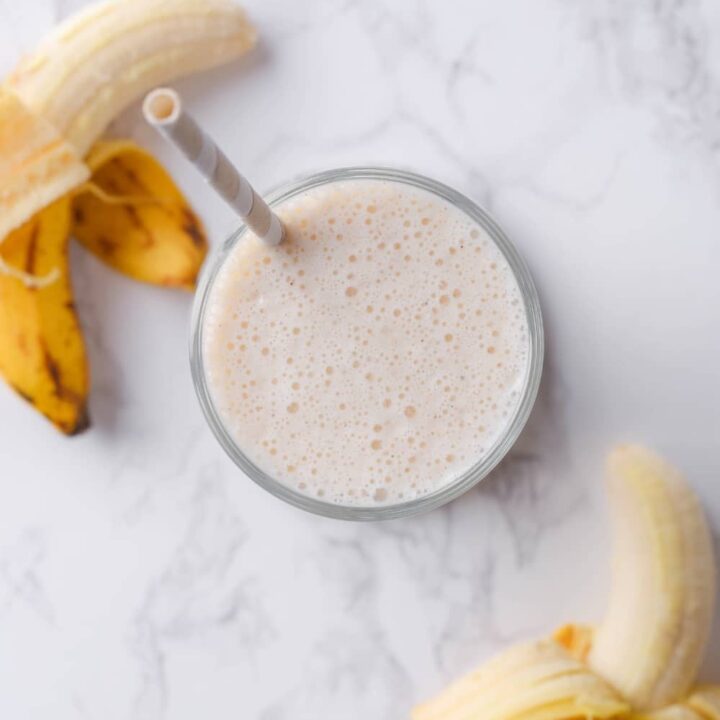 A banana protein shake in a glass on a counter with two bananas next to it.