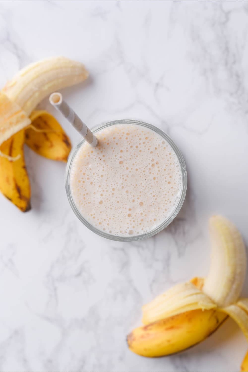 A banana protein shake in a glass on a counter with two bananas next to it.