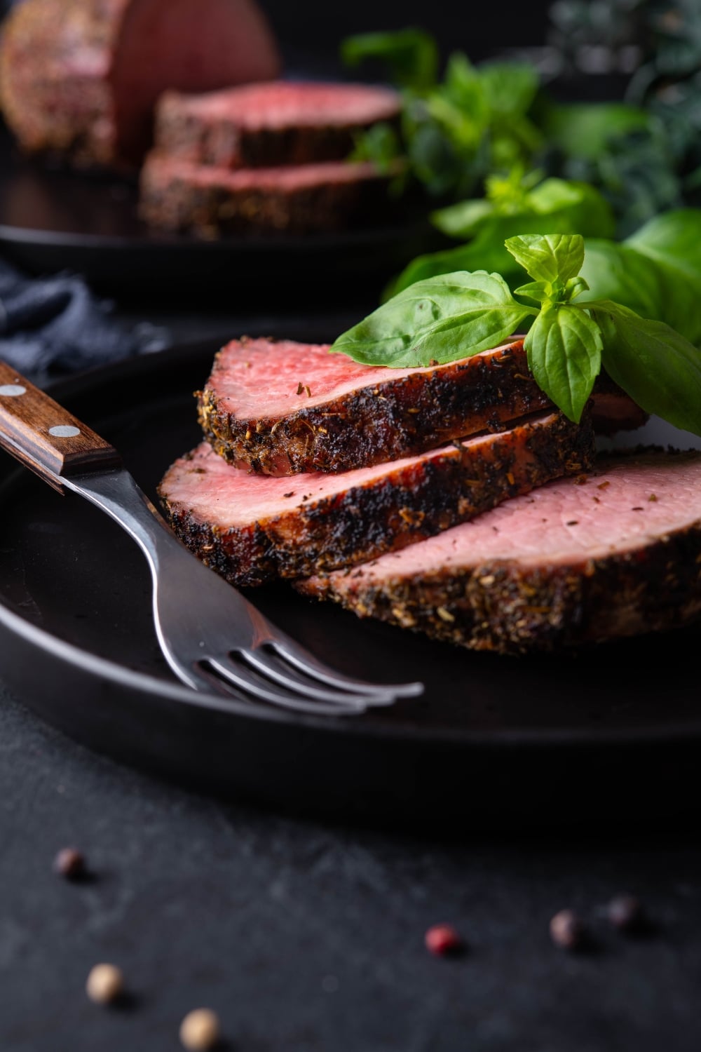 Sliced medium rare grilled beef tenderloin on a black plate, garnished with basil and served with a fork. In the back is another plate with the rest of the grilled beef tenderloin.