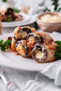 Four pieces of eggplant rollatini on a white plate garnished with curly parsley. Behind is a second plate of eggplant rollatini and a bowl of shredded cheese.