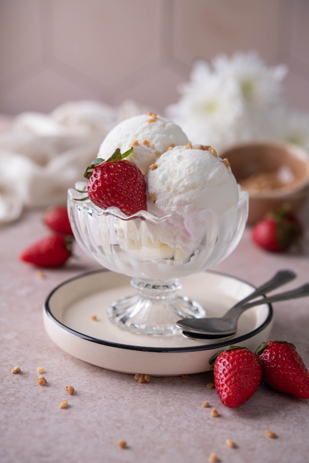 Scoops of frozen yogurt ice cream served in a glass ice cream bowl over a plate, garnished with strabwerries and ground hazelnut brittle.