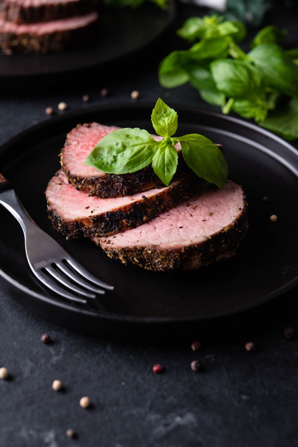 Three slices of medium rare grilled beef tenderloin on a black plate, garnished with basil and served with a fork.