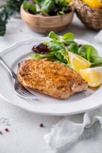 A grilled mahi mahi filet on a plate with salad greens and lemon wedges. Behind is a small bowl with more salad greens.