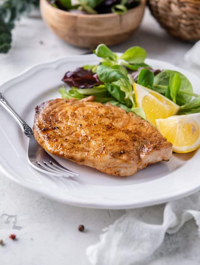 A grilled mahi mahi filet on a plate with salad greens and lemon wedges. Behind is a small bowl with more salad greens.