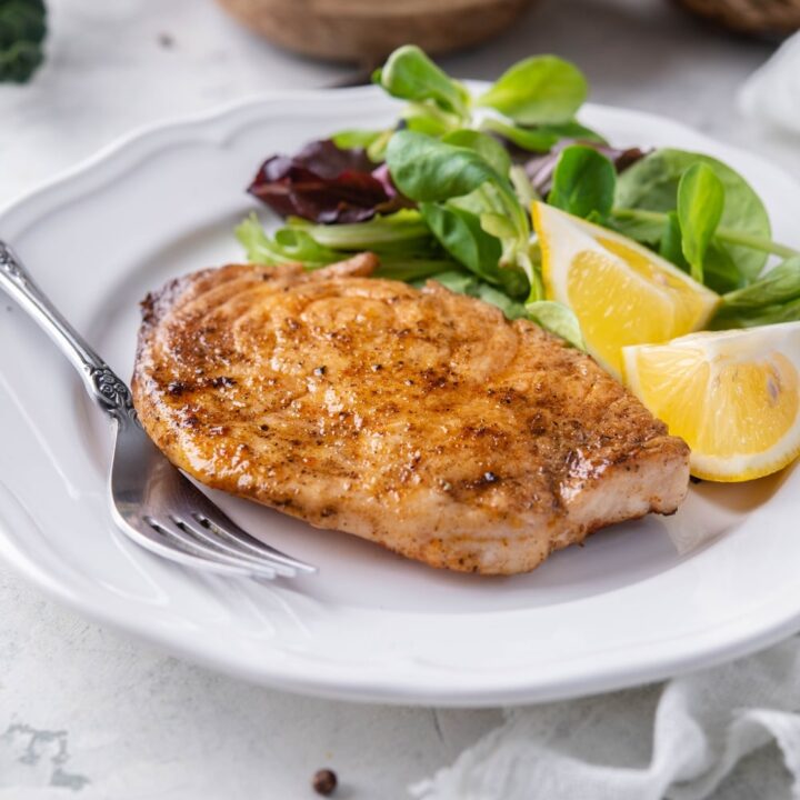 A grilled mahi mahi filet on a plate with salad greens and lemon wedges. Behind is a small bowl with more salad greens.