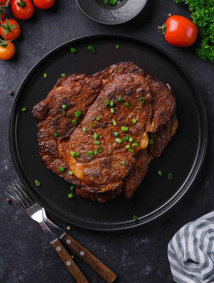 Top view of grilled ribeye steaks on a black plate garnished with spring onions.