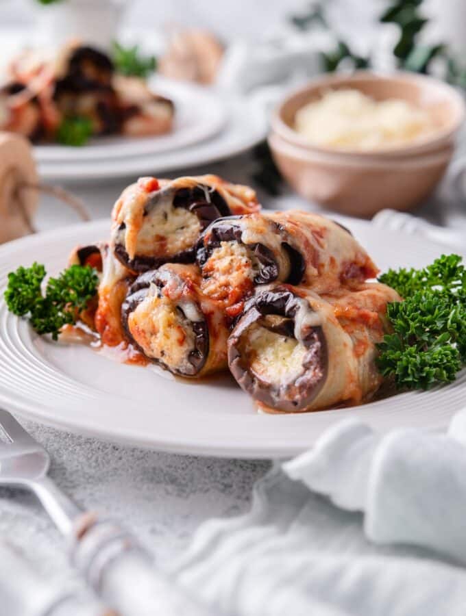 Four pieces of eggplant rollatini garnished with curly parsley. A second plate of eggplant rollatini and a bowl of shredded cheese are in the back.