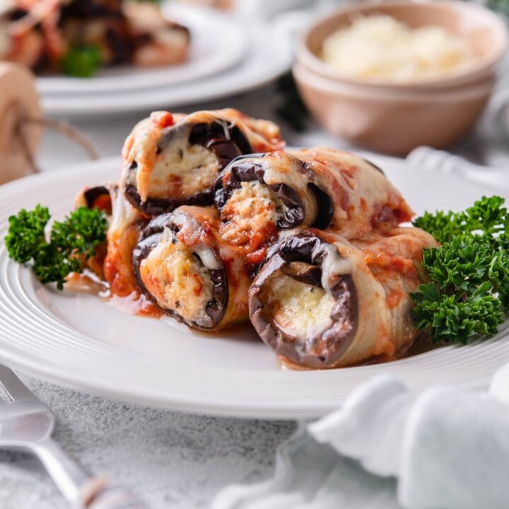 Four pieces of eggplant rollatini garnished with curly parsley. A second plate of eggplant rollatini and a bowl of shredded cheese are in the back.