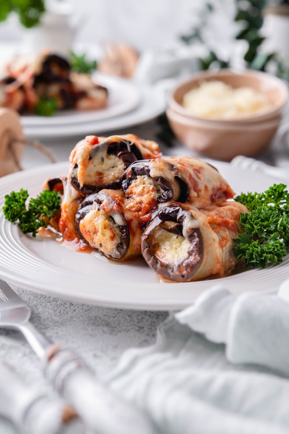 Four pieces of eggplant rollatini garnished with curly parsley. A second plate of eggplant rollatini and a bowl of shredded cheese are in the back.