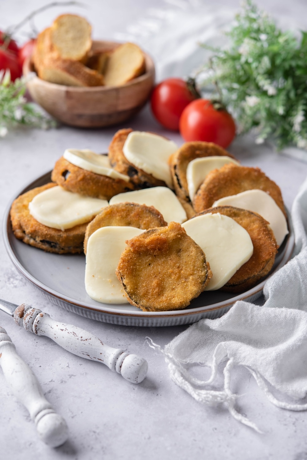 Fried eggplant slices layered with slices of mozzarella cheese on a plate. In the back is a bowl of bread slices and fresh tomatoes.