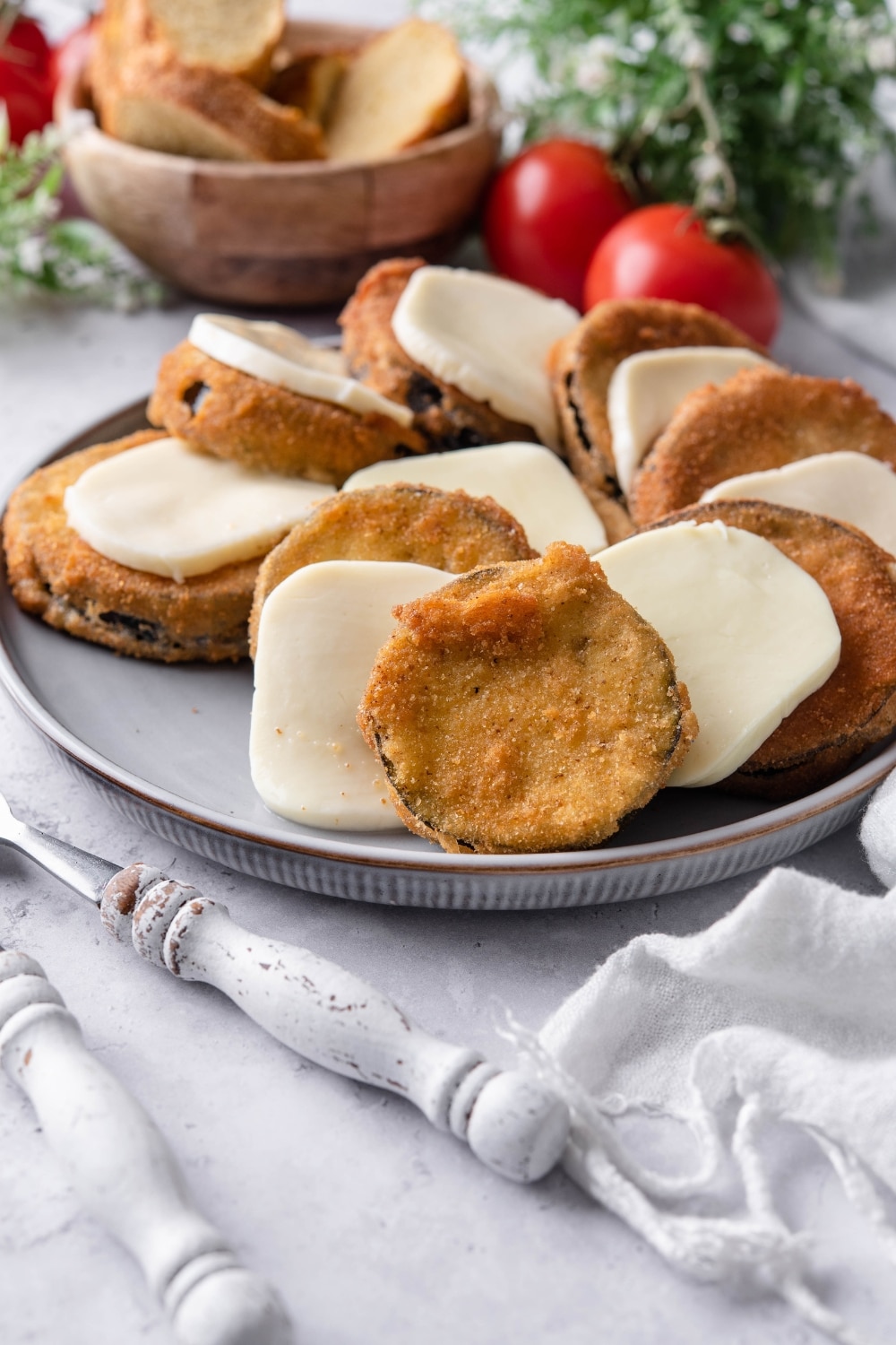 Fried eggplant slices with mozzarella cheese on a plate in front of a bowl of bread and fresh tomatoes.