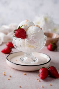 Frozen yogurt ice cream scoops, garnished with a strawberry and ground hazelnut brittle, served in a glass ice cream bowl over a plate.