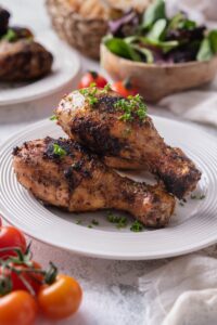 Grilled chicken drumsticks on a white plate, garnished with parsley. In the back is a bowl of salad, fresh cherry tomatoes, and another plate of grilled chicken drumsticks.