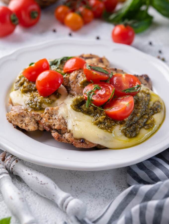 Grilled chicken margherita topped with mozzarella, pesto, and cherry tomatoes on a white plate.