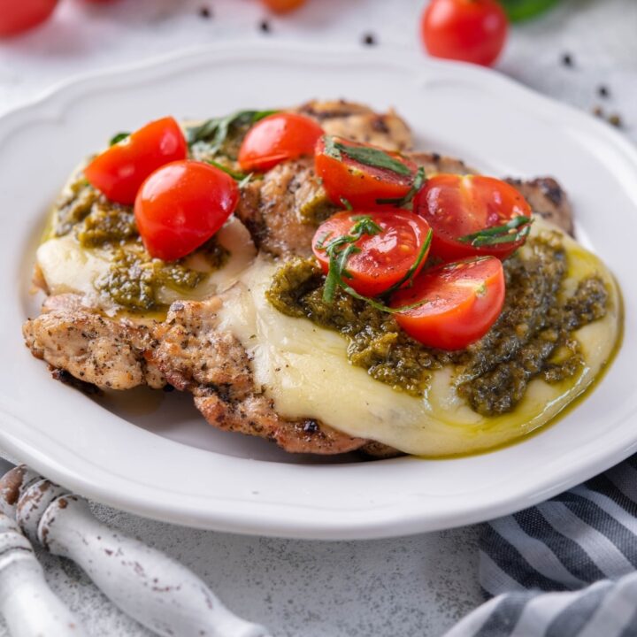 Grilled chicken margherita topped with mozzarella, pesto, and cherry tomatoes on a white plate.