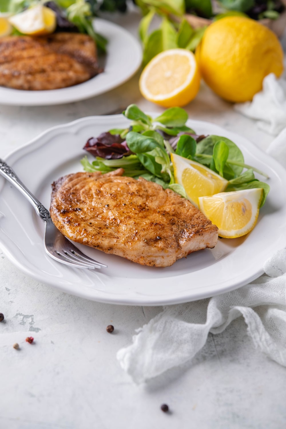 A grilled mahi mahi filet on a plate with salad greens and lemon wedges. Behind is a second plate of grilled mahi mahi with salad.