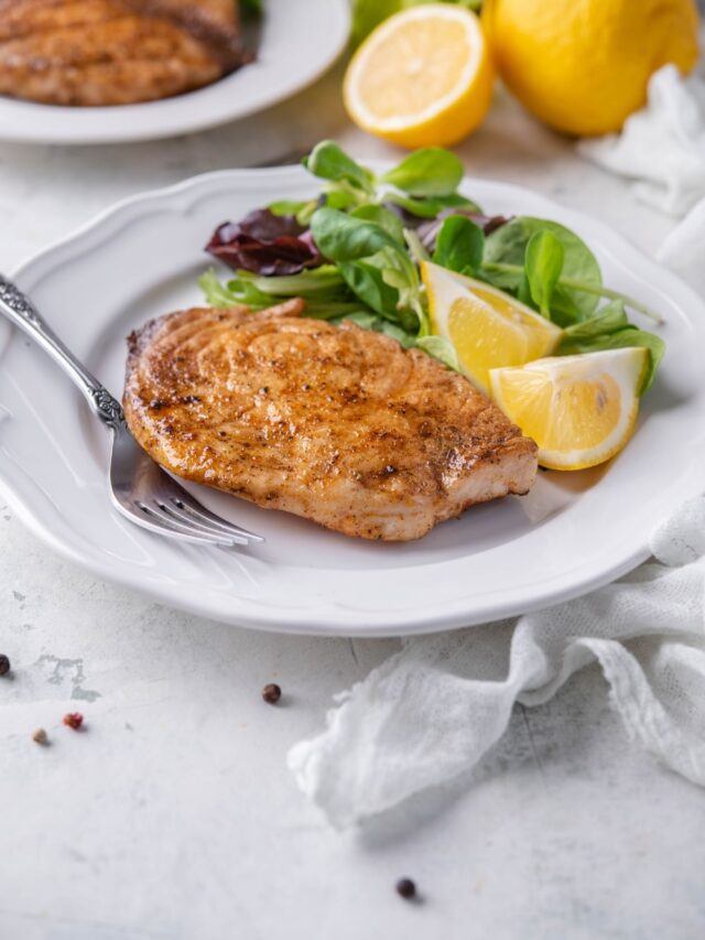 A grilled mahi mahi filet on a plate with salad greens and lemon wedges. Behind is a second plate of grilled mahi mahi with salad.