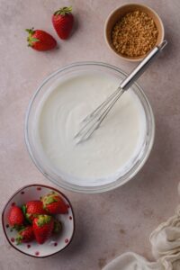 Greek yogurt and sugar mixture in a glass bowl with a whisk, next to small bowls of fresh strawberries and ground hazelnut brittle.