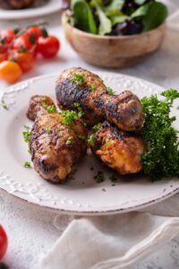 Grilled chicken drumsticks garnished with parsley on a white plate. Behind them is a bowl of salad and fresh cherry tomatoes.