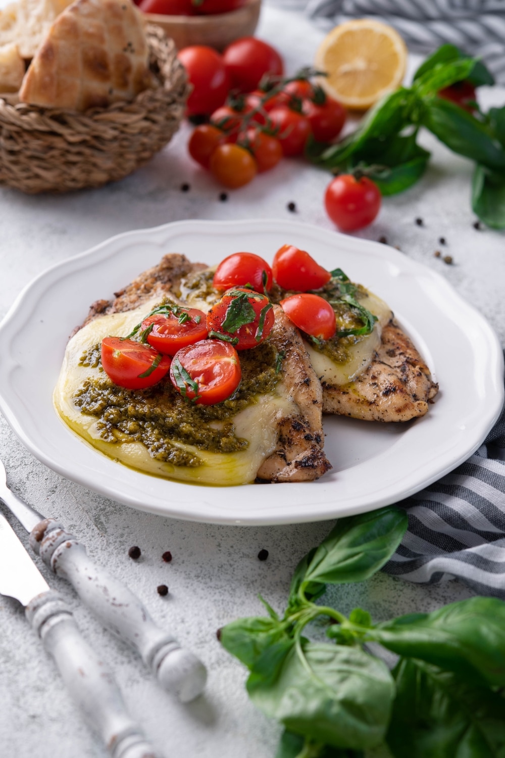 Two pieces of herb grilled chicken topped with mozzarella, pesto, and cherry tomatoes on a white plate.