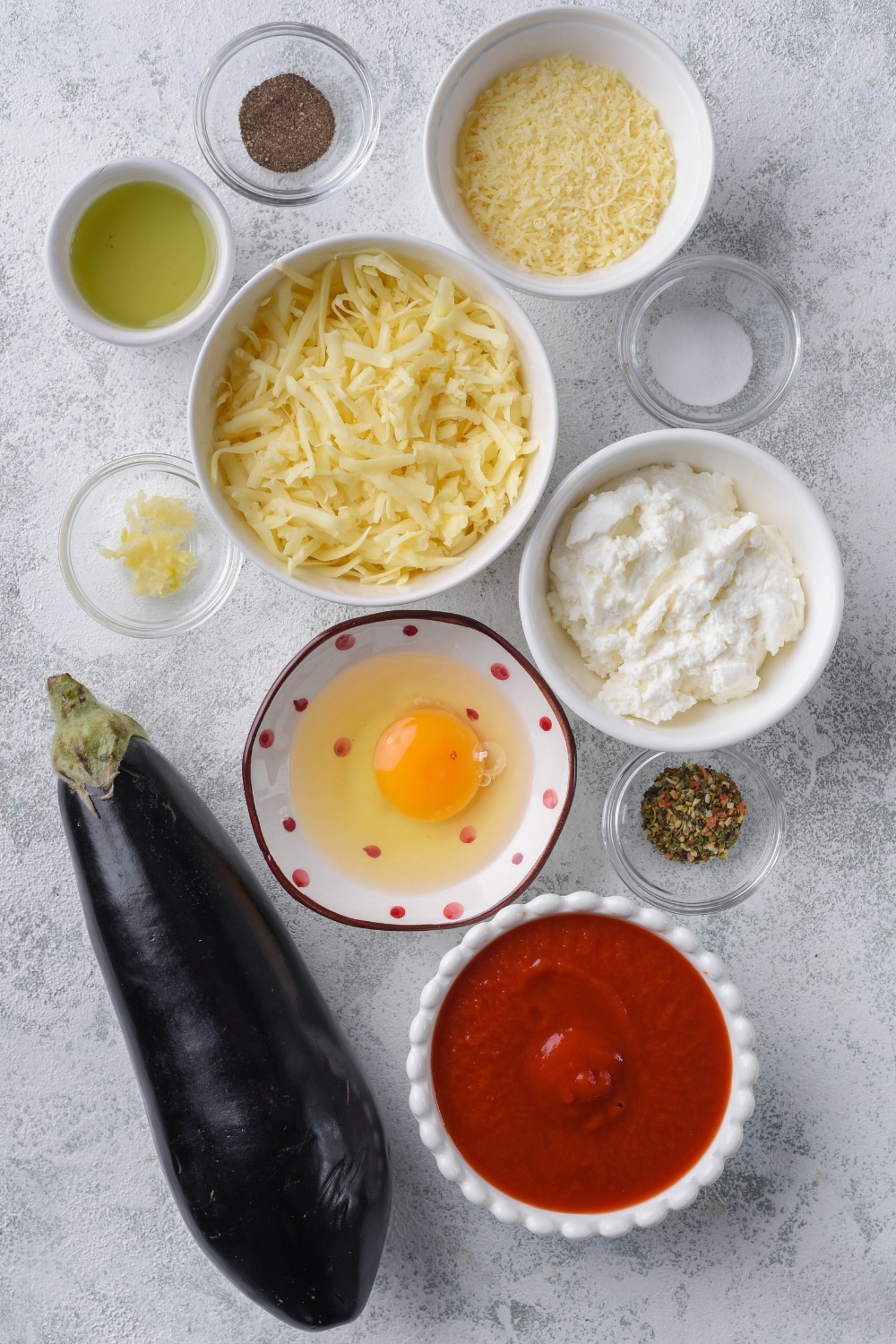 A large eggplant and bowls of ricotta cheese, shredded mozzarella cheese, tomato sauce, egg, olive oil, and spices.