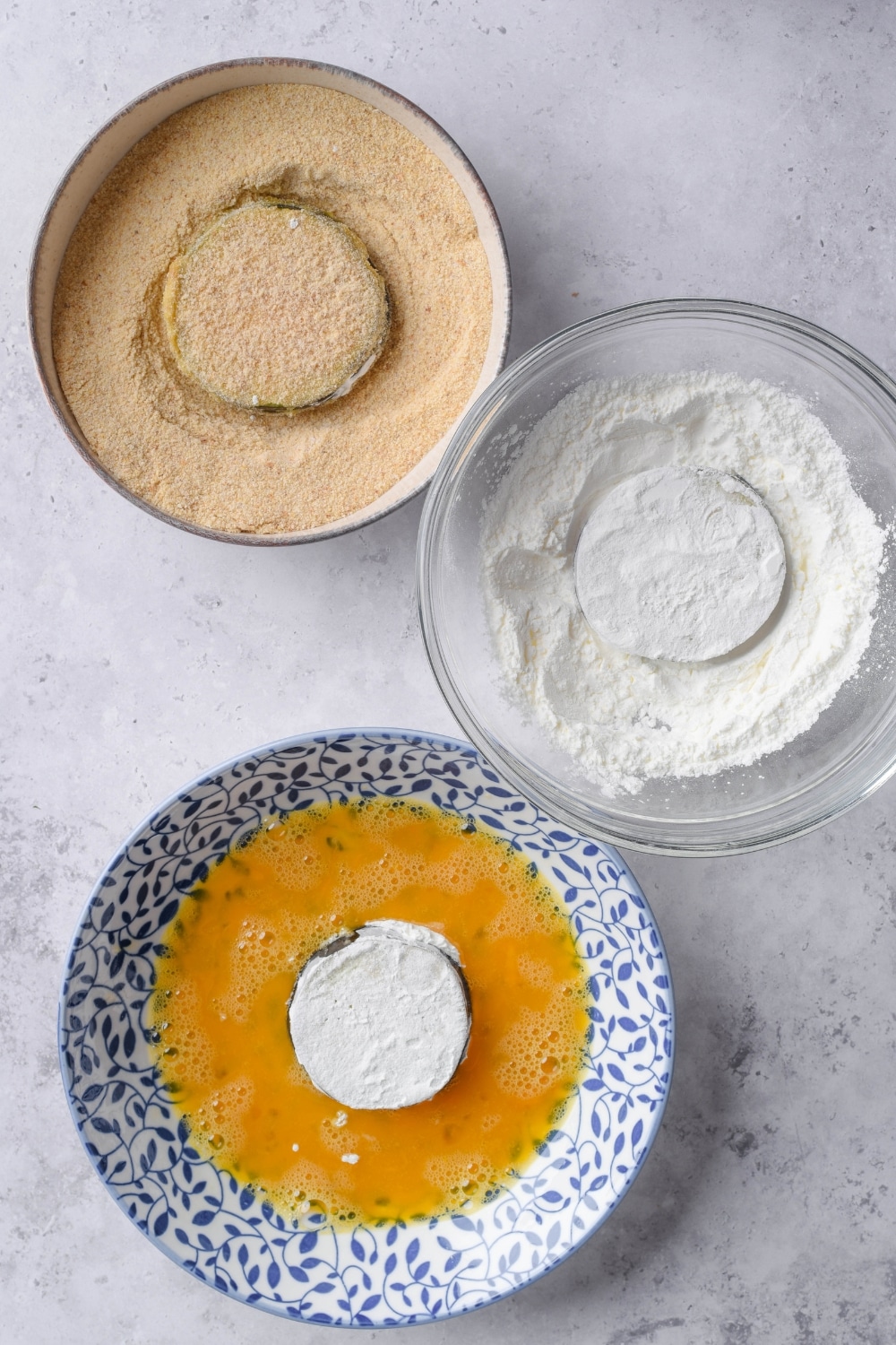 Bowls of breadcrumbs, cornstarch, and egg wash, each with an uncooked breaded eggplant slice in the center.