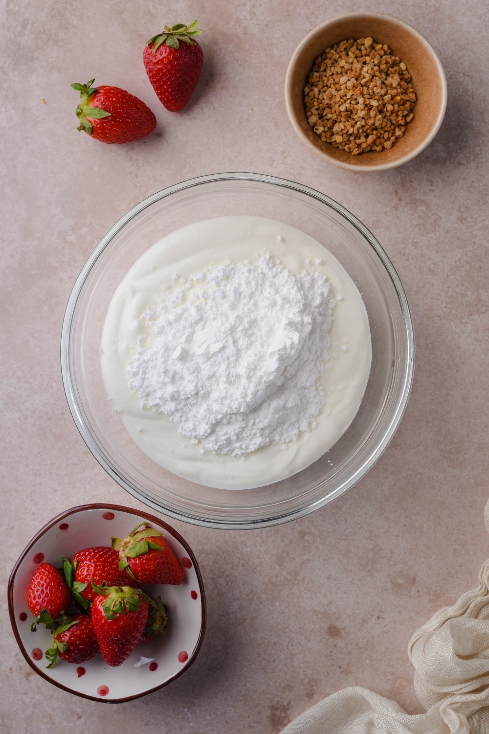 Unmixed greek yogurt and powdered sugar in a glass bowl, next to small bowls of fresh strawberries and crushed hazelnut brittle.