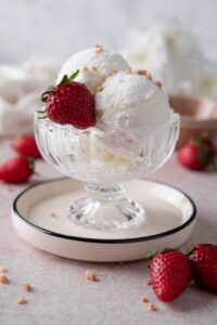 Scoops of yogurt ice cream, garnished with a fresh strawberry and ground hazelnut brittle in a small ice cream bowl.