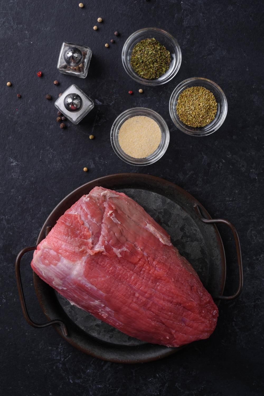 Raw beef tenderloin on a cast iron plate with small glass bowls of dried thyme, dried rosemary, garlic powder, and salt and pepper shakers.