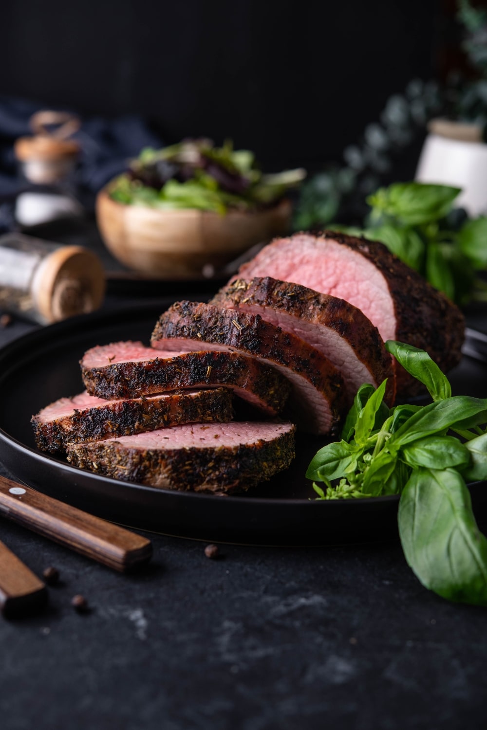 Sliced whole grilled beef tenderloin on black plate with fresh basil. A bowl of salad greens is in the back.