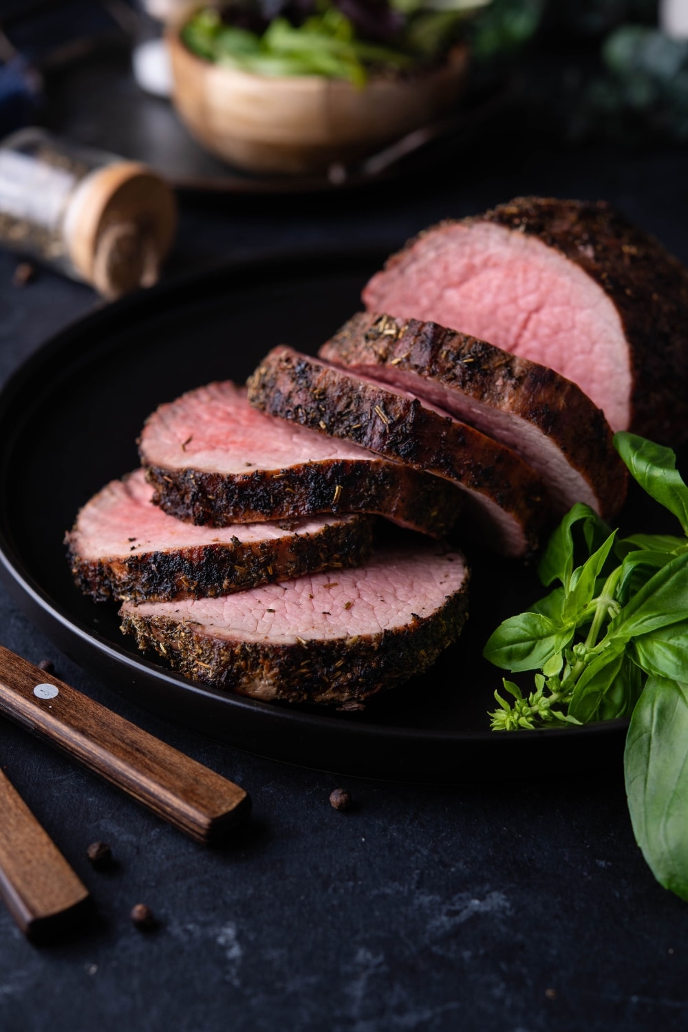Medium rare grilled beef tenderloin, served and served on a black plate with basil leaves.