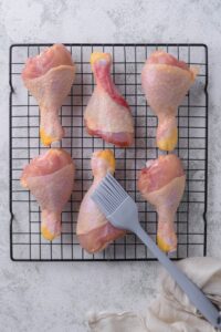 Six raw chicken drumsticks on a drying rack with a pastry brush.