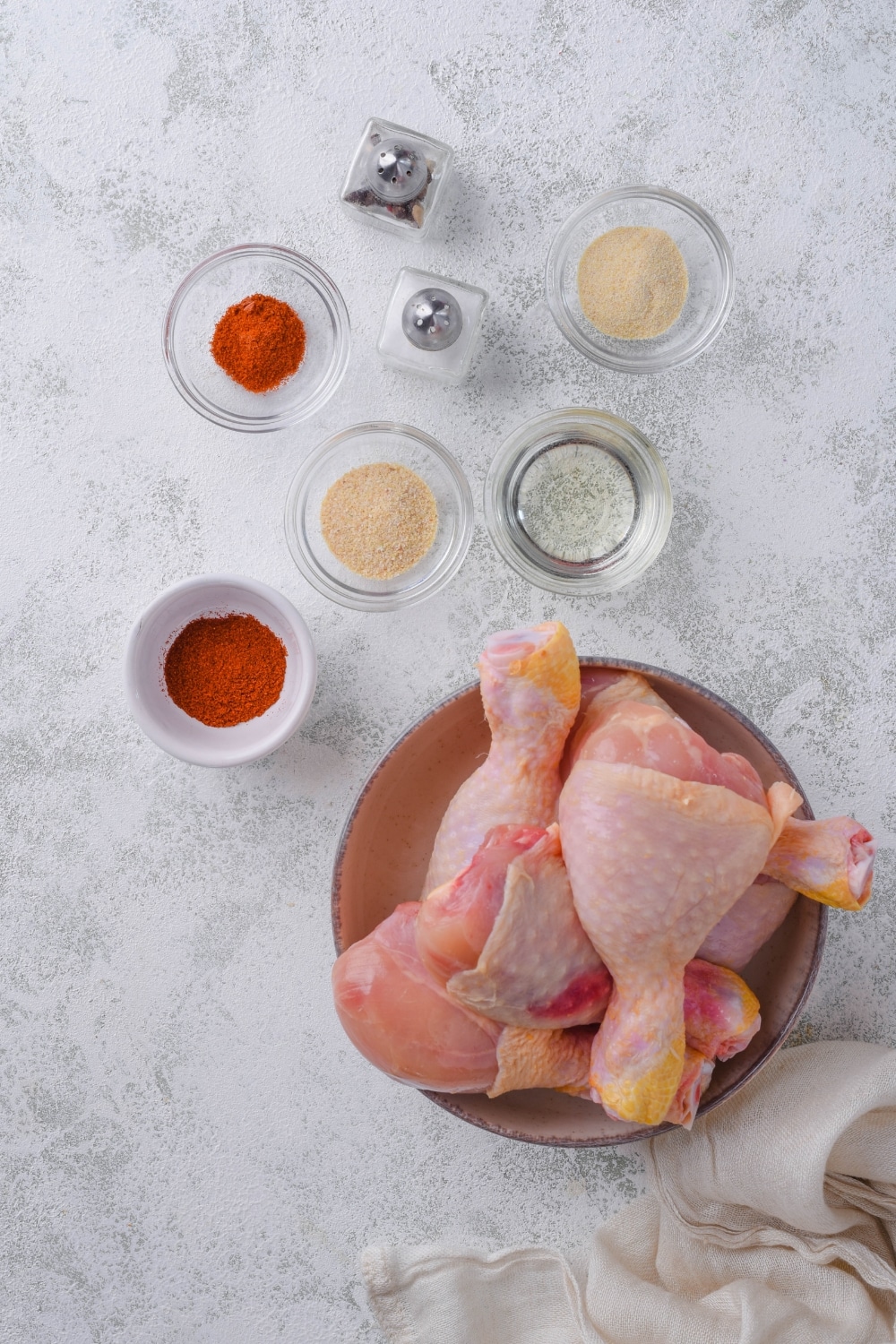 A bowl of raw chicken legs, small bowls of spices, and salt and pepper shakers.