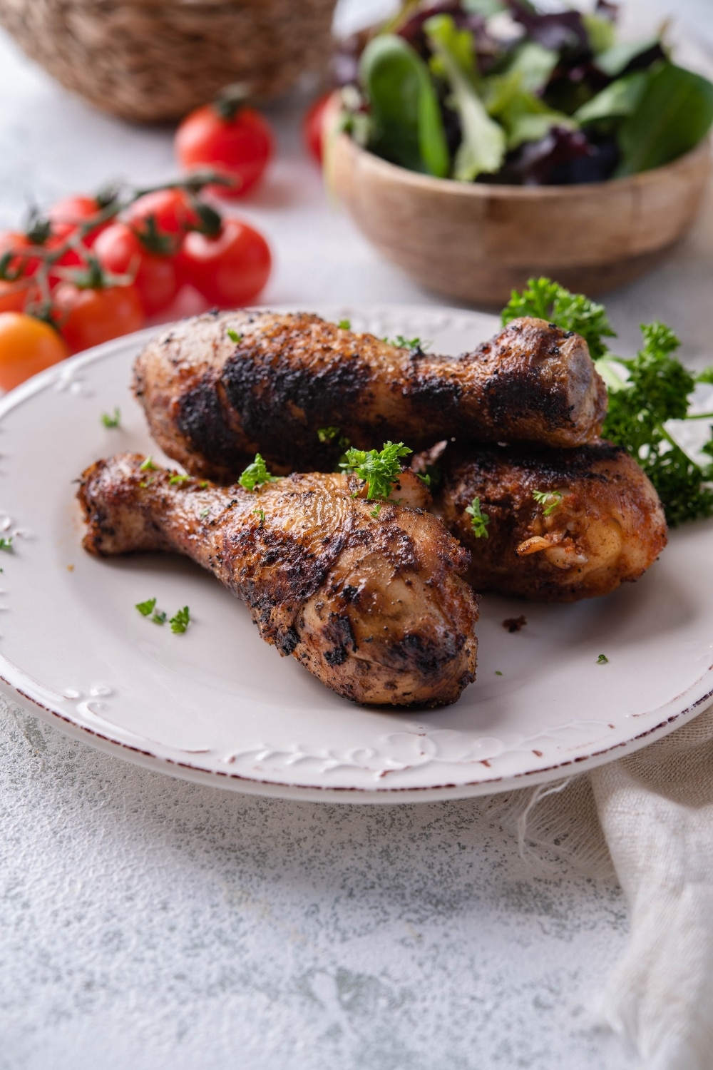 Three grilled chicken legs garnished with parsley on a white plate. Behind them is a bowl of salad and fresh cherry tomatoes.