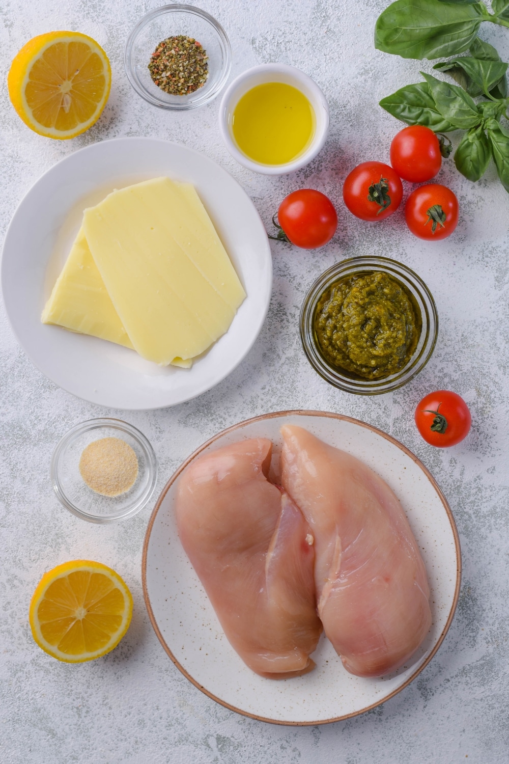 A plate of two raw chicken breast filets, a plate of mozzarella slices, small bowls of pesto, italian seasoning, oil, and garlic powder, a halved lemon, and scattered cherry tomatoes.