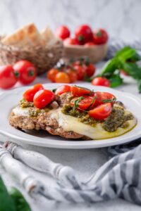 Two pieces of grilled chicken margherita served on a white plate. In the back are bowls of bread and tomatoes.