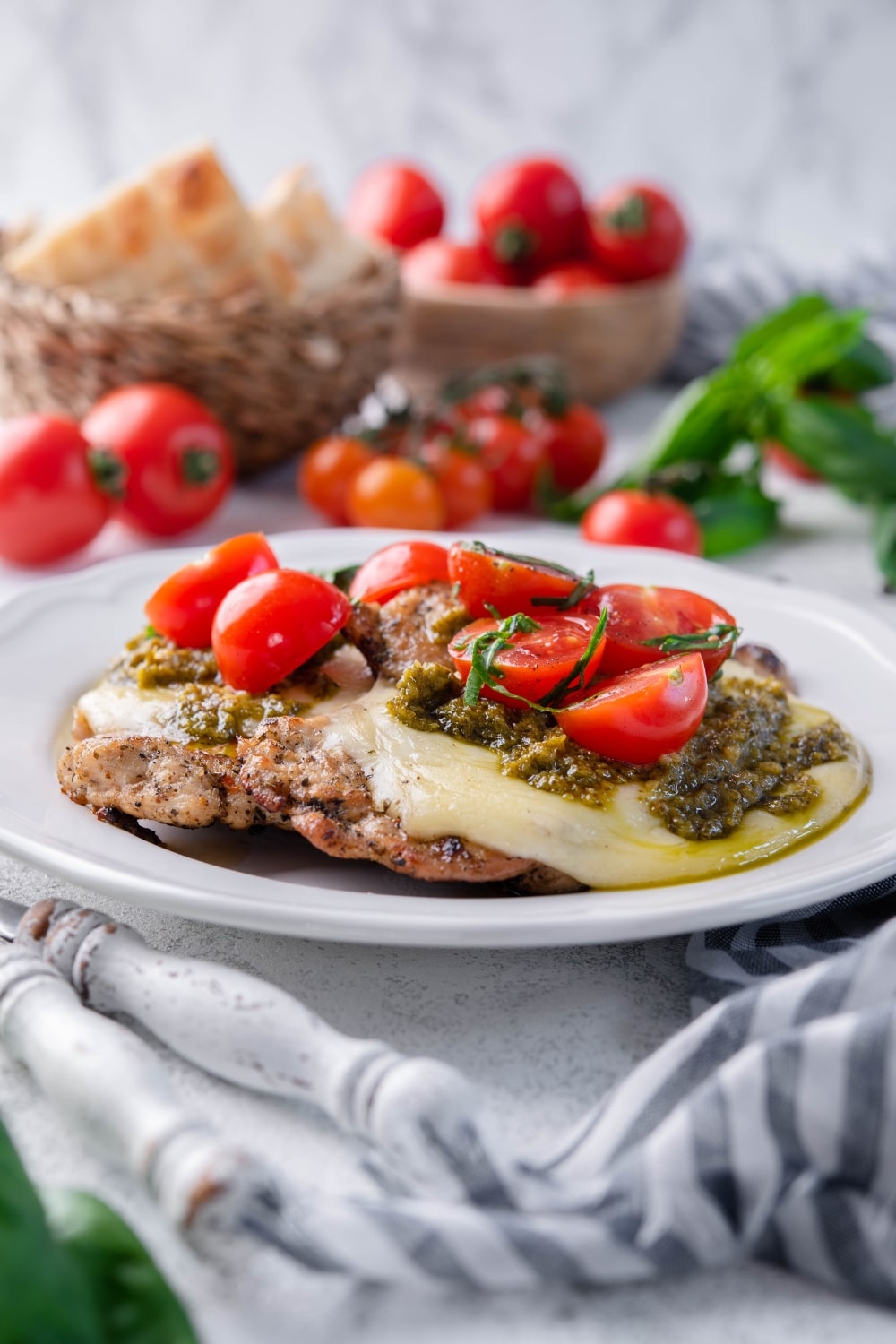 Two pieces of grilled chicken margherita served on a white plate. In the back are bowls of bread and tomatoes.