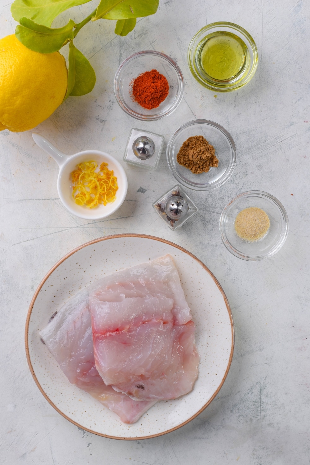 A plate of raw halibut filets, small glass bowls of spices and olive oil, and salt and pepper shakers.