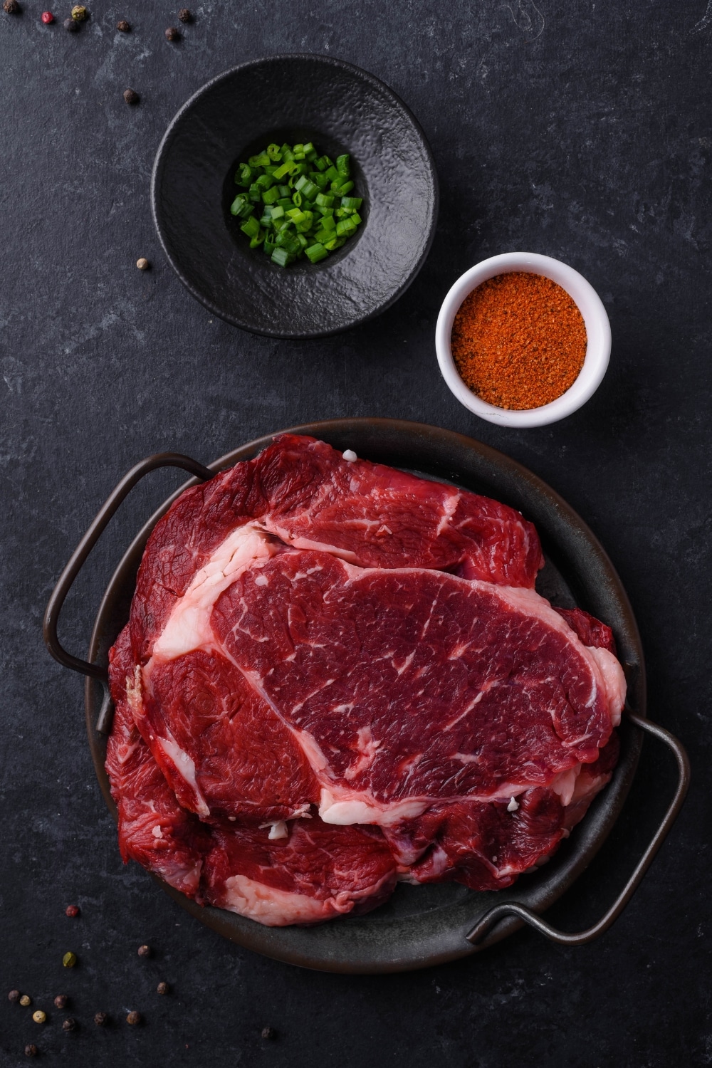 Raw ribeye steaks on a cast iron plate, a small bowl of dry rub, and chopped spring onions on a small black plate.