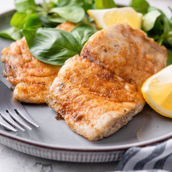 Three pieces of grilled halibut on a plate with lemon wedges and lettuce.