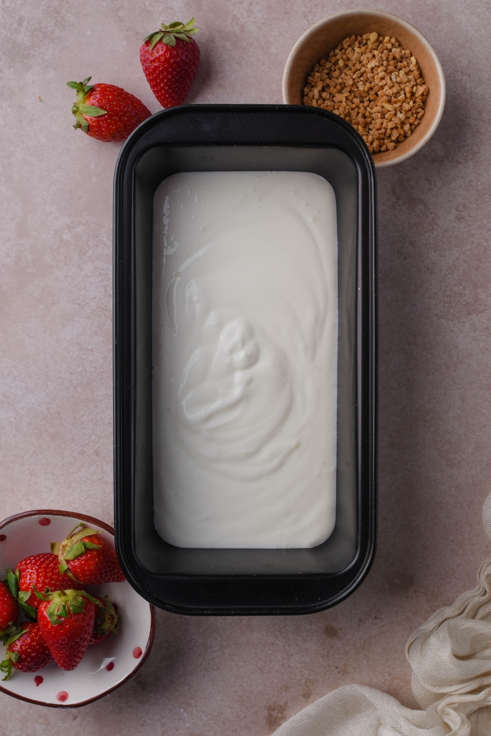 Yogurt mixture in a freezer-safe container next to bowls of ground hazelnut brittle and fresh strawberries.